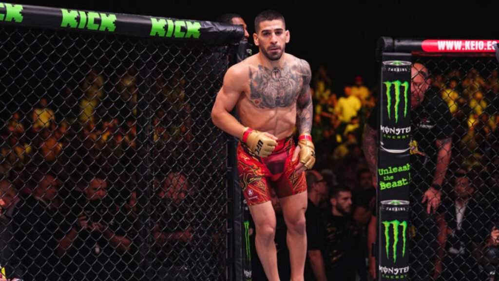 A tattooed Ilia Topuria stands focused inside the UFC octagon, wearing red and gold fight shorts.