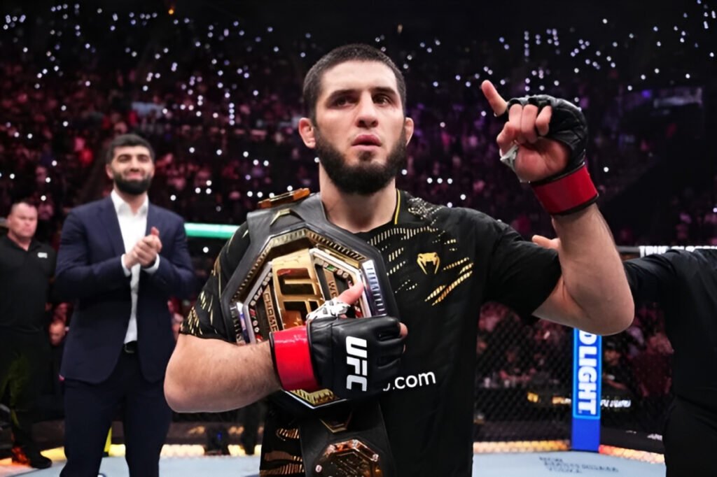 UFC champion Islam Makhachev holds his title belt in the octagon, raising one finger to the crowd in victory.