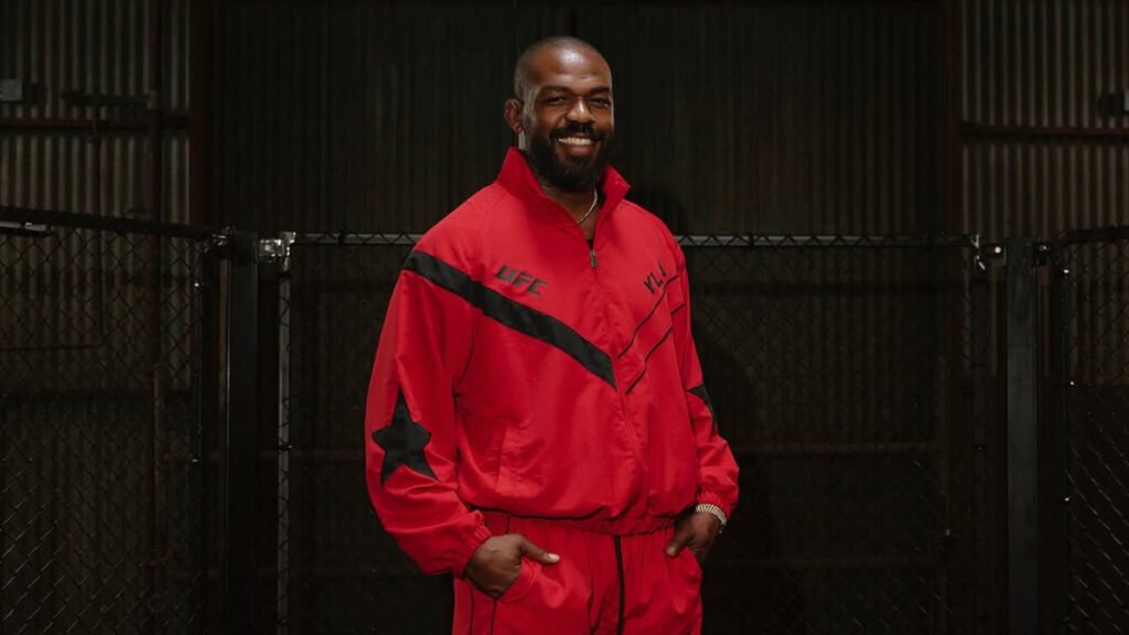 El luchador de la UFC, Jon Jones, sonríe con un chándal rojo y negro frente a una jaula de combate.