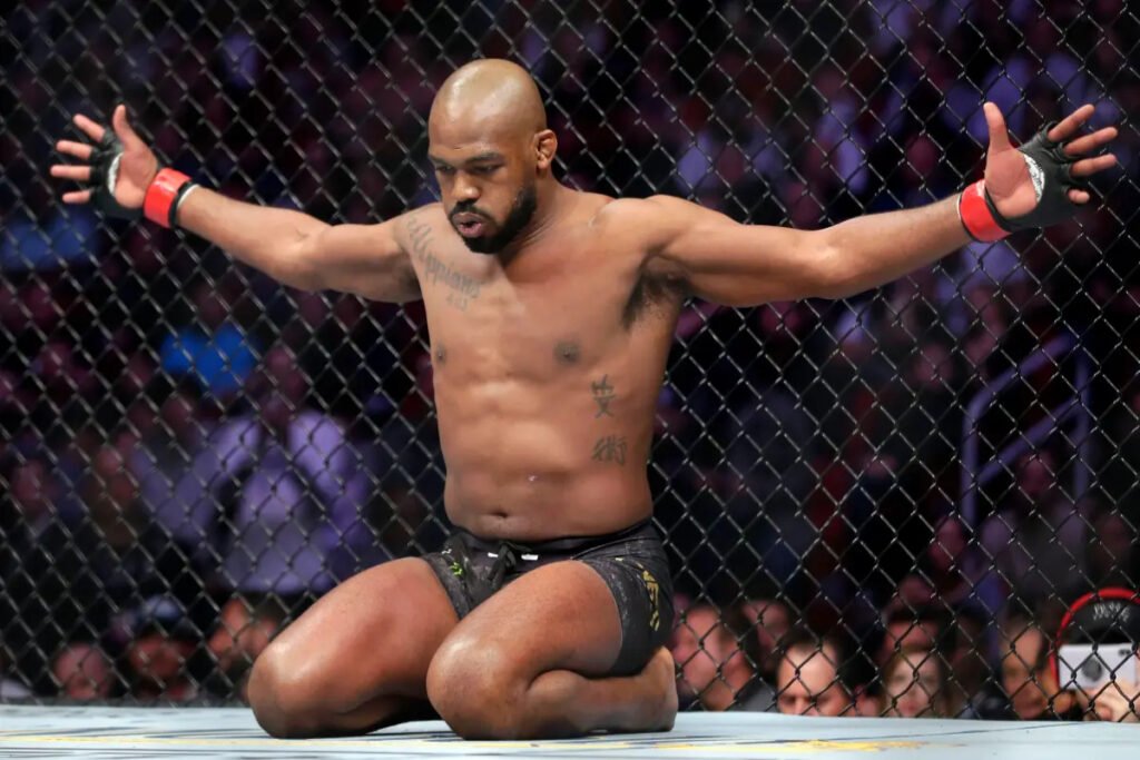 UFC fighter Jon Jones kneels on the mat inside the octagon with his arms spread wide.