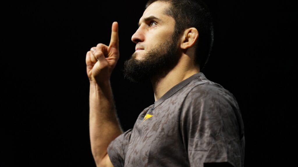 A profile shot of bearded fighter Islam Makhachev pointing his index finger upward against a black background.