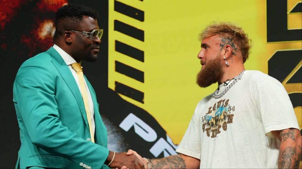 Francis Ngannou, con traje verde, y Jake Paul, con barba y tatuajes, se dan la mano en un cara a cara.