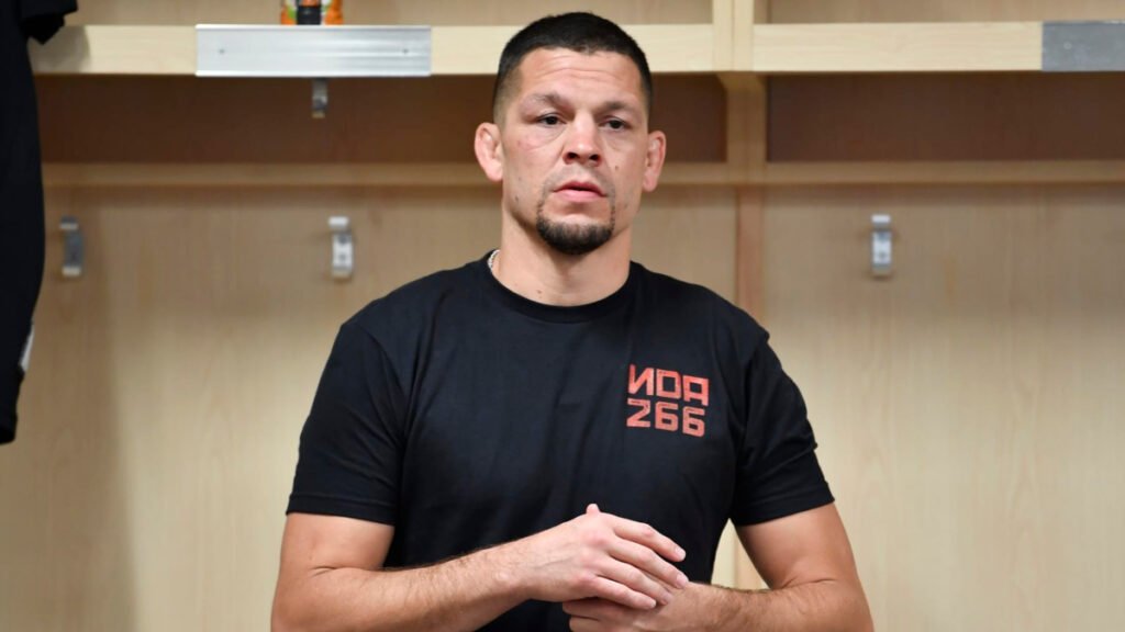 MMA fighter Nate Diaz stands in a locker room with a serious expression, wearing a black graphic t-shirt.
