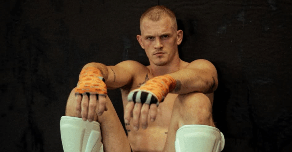 Luchador de MMA con pelo rapado, vendas y espinilleras, sentado con una mirada intensa.