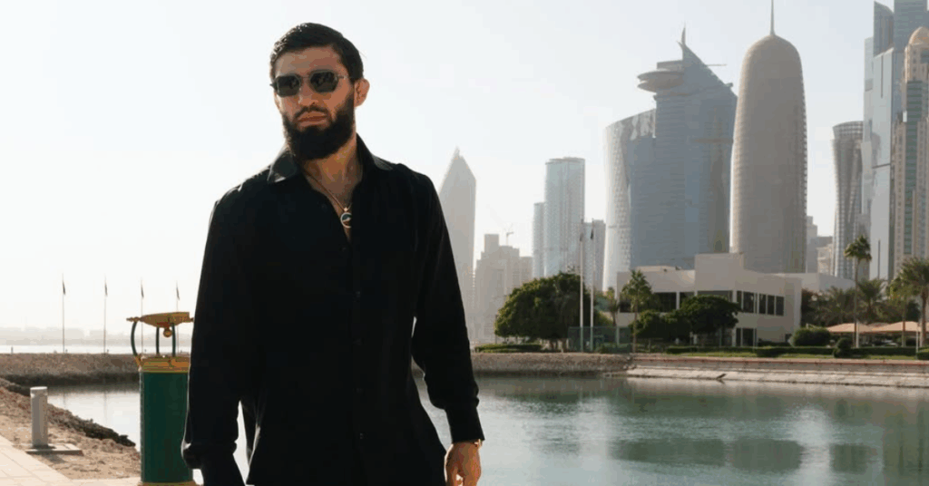Hombre barbudo con gafas de sol y camisa negra posando frente al moderno horizonte de rascacielos de Doha, Catar.