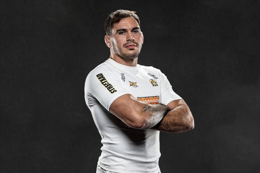 A muscular fighter with short brown hair poses with his arms crossed in a white shirt against a dark studio background.