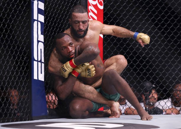 UFC fighters Belal Muhammad and Leon Edwards grapple against the cage, with Muhammad on Edwards' back.