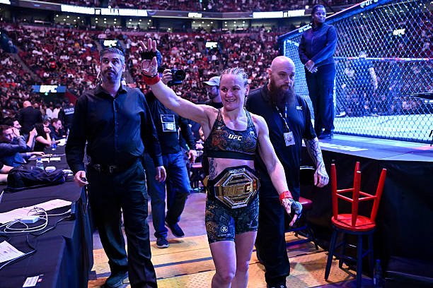 A smiling female UFC champion raises her hand in victory, wearing her title belt as she walks in a crowded arena.
