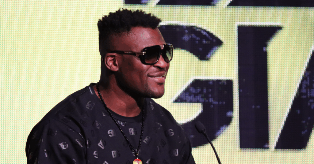 El luchador Francis Ngannou sonríe de perfil, usando gafas de sol y una camiseta negra estampada frente a un micrófono.