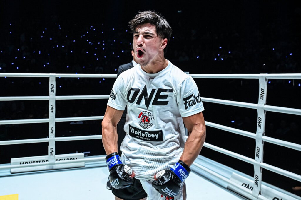 Luchador de ONE Championship celebra en el ring, gritando con guantes Fairtex. Camiseta Tiger Muay Thai, público oscuro con luces azules.