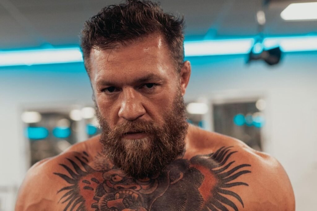 Conor McGregor, luchador barbudo y musculoso, con tatuajes en el pecho y mirada intensa. Fondo azulado difuminado.