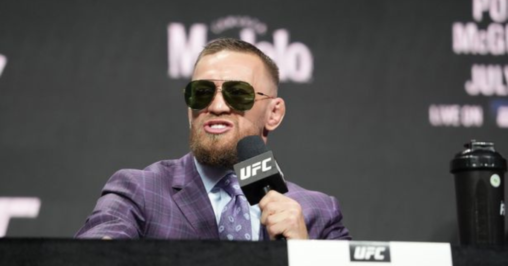 Conor McGregor, con gafas de sol y traje a cuadros morado, discursa en rueda de prensa de UFC, micrófono en mano.