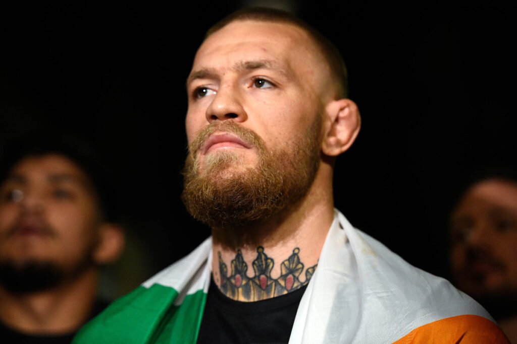 Conor McGregor, luchador de UFC, con barba, bandera irlandesa al cuello y tatuaje de coronas, mira fijamente hacia arriba.