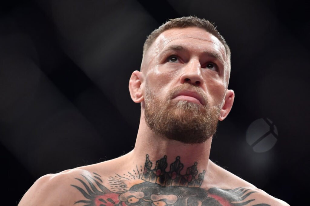 Conor McGregor, estrella de UFC, mira con concentración y gesto serio. Rostro sudoroso, barba y tatuajes en el octágono oscuro.