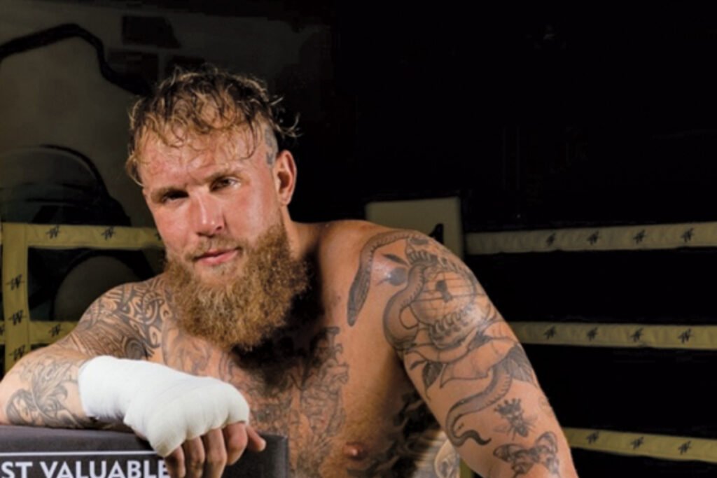 Boxeador barbudo y tatuado, con vendas blancas en la mano, mira intenso desde un ring de boxeo. Fondo oscuro.