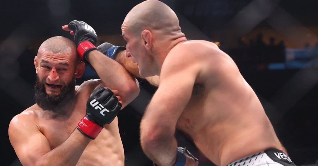 Luchador de MMA barbudo con nariz sangrienta y expresión de dolor tras un golpe, mientras su oponente calvo lucha cerca en el octágono.