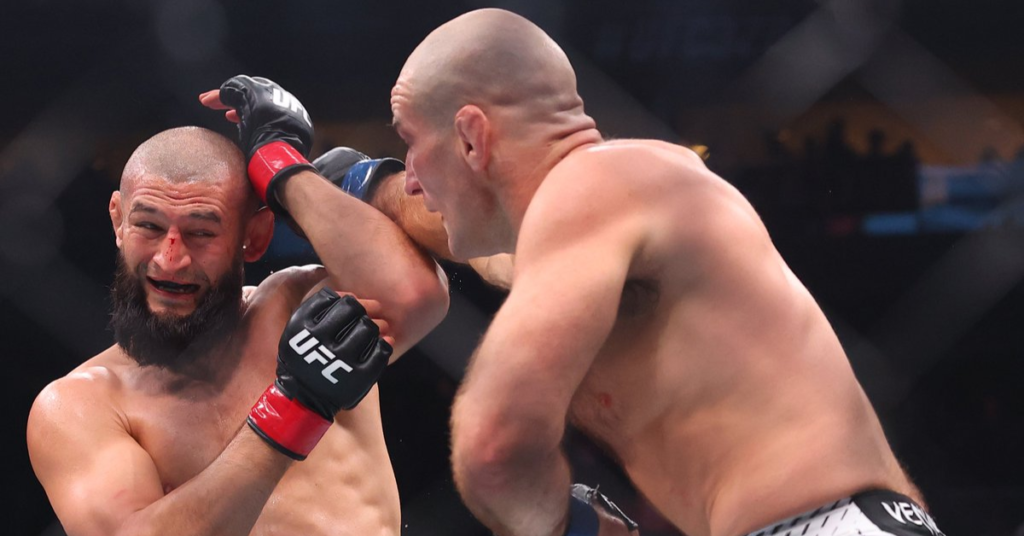 Combate de UFC: un luchador barbudo con nariz sangrando reacciona a un golpe de su rival calvo en la jaula.