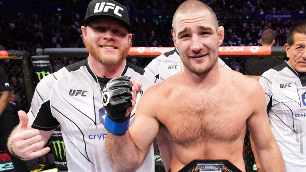 El campeón de UFC Sean Strickland sonríe con su cinturón. A su lado, un hombre con gorra y guante de MMA celebra con un pulgar arriba.