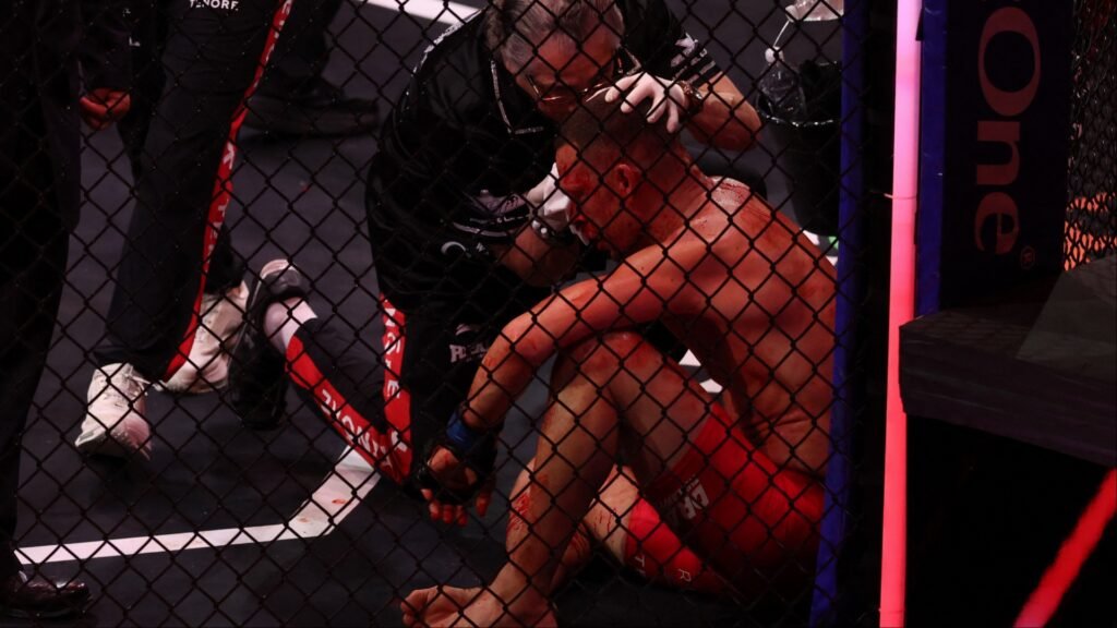 Luchador de MMA ensangrentado, sentado en la jaula, recibe asistencia médica en la cabeza.