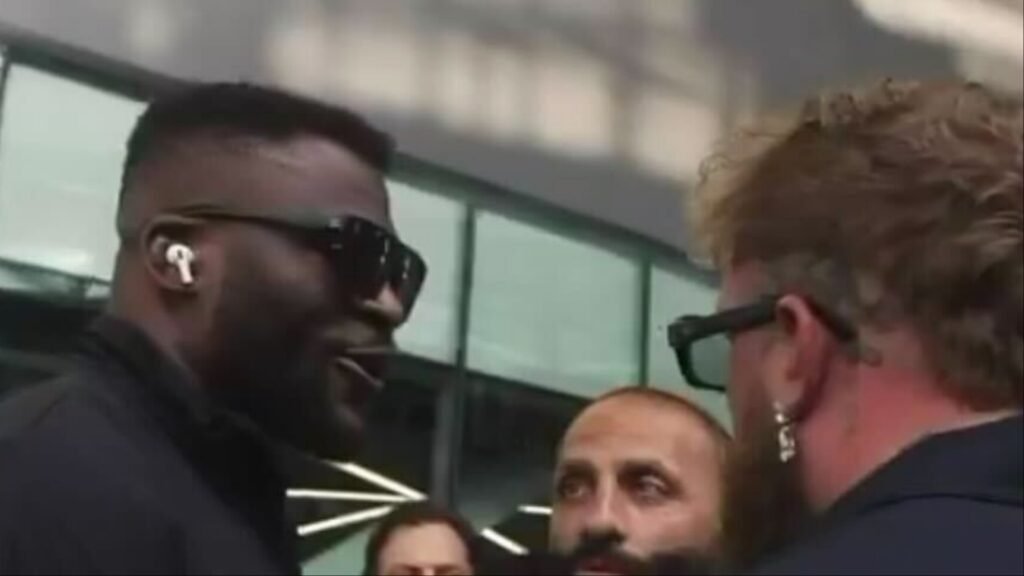 Intensa confrontación cara a cara entre dos hombres; uno negro con AirPods y gafas de sol, otro con pelo rizado y pendiente. Un mediador barbudo.
