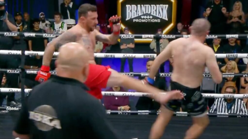 Combate en ring: un luchador con guante rojo patea a su oponente. Árbitro ISKA de espaldas. Público y cartel BRANDRISK PROMOTIONS.