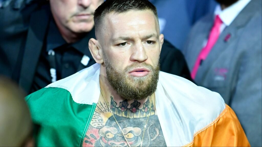 Conor McGregor, luchador de UFC, envuelto en la bandera irlandesa. Rostro serio con barba y tatuajes en el cuello.