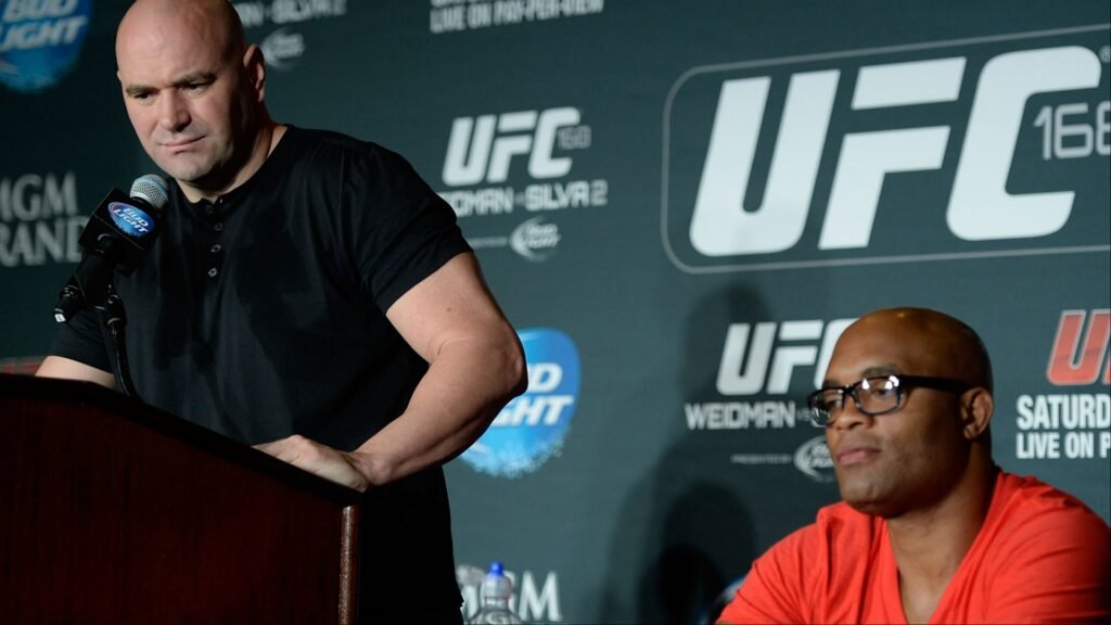 Dana White en un podio de UFC 168. Anderson Silva, luchador de MMA, sentado, con logos UFC de fondo.