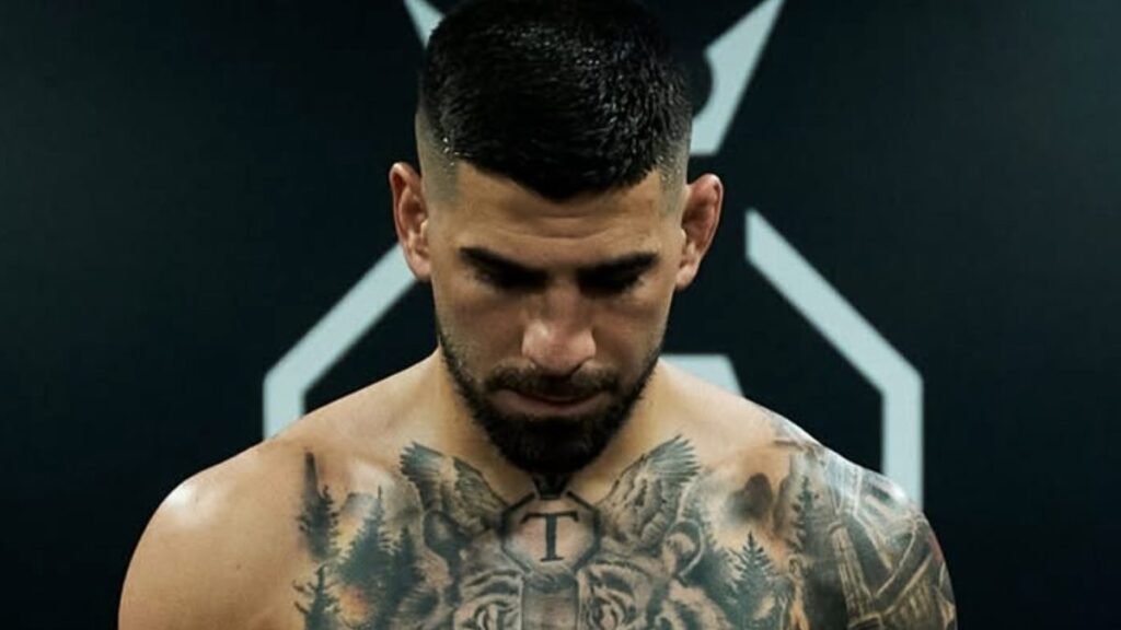 Hombre con barba y pelo corto, mirando hacia abajo. Destacan sus tatuajes de tigre, una 'T' y árboles en el pecho y brazo.