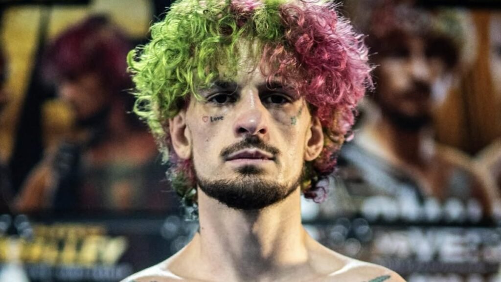 El luchador Sean O'Malley de UFC, con su icónico pelo rizado verde y rosa, tatuajes faciales y mirada penetrante.
