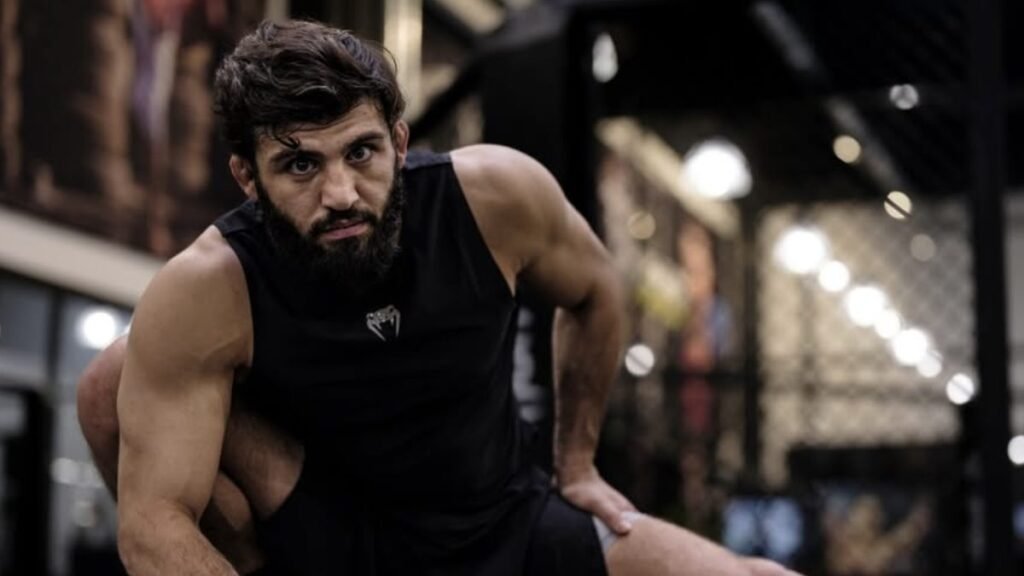 Luchador barbudo con camiseta Venum negra, mirada intensa, estirándose en un gimnasio.