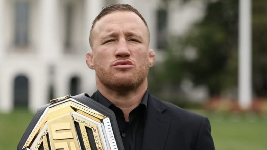 Luchador de UFC sosteniendo cinturón de campeón. Hombre con barba, orejas de coliflor y traje oscuro, fondo desenfocado.