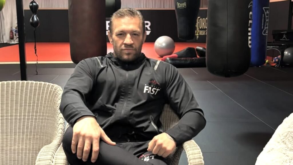 Atleta en chándal negro con logo FAST, sentado en silla de mimbre. De fondo, gimnasio con sacos de boxeo y equipamiento deportivo.