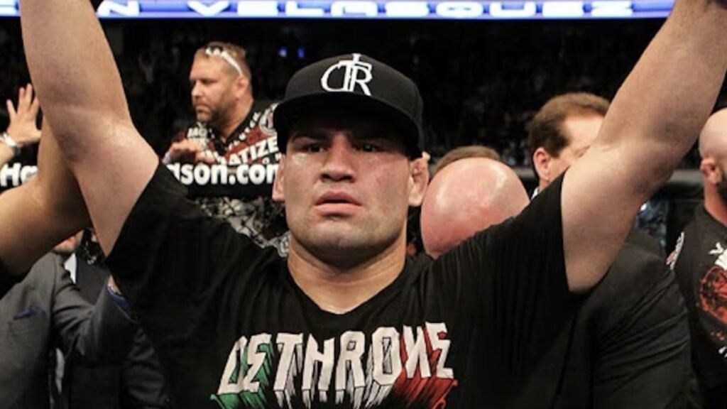 Cain Velasquez, campeón de MMA, celebra una victoria con brazos levantados. Luce gorra y camiseta 'DETHRONE' en la arena.