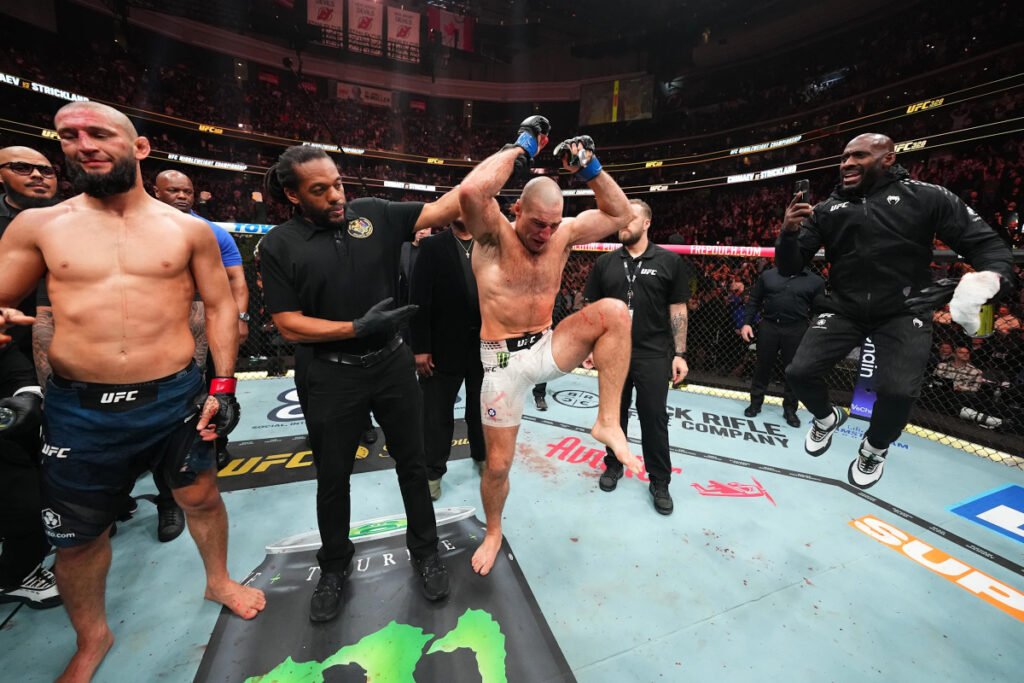 Sean Strickland celebra su victoria eufórico en el octágono de UFC. Chimaev, herido, árbitro y eufórico entrenador acompañan.