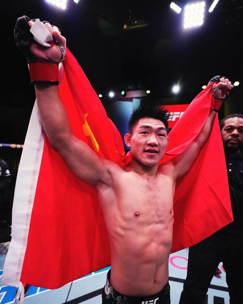 Luchador Song Yadong celebra con bandera china en el octágono de UFC tras su victoria. Brazos en alto, mirada satisfecha.