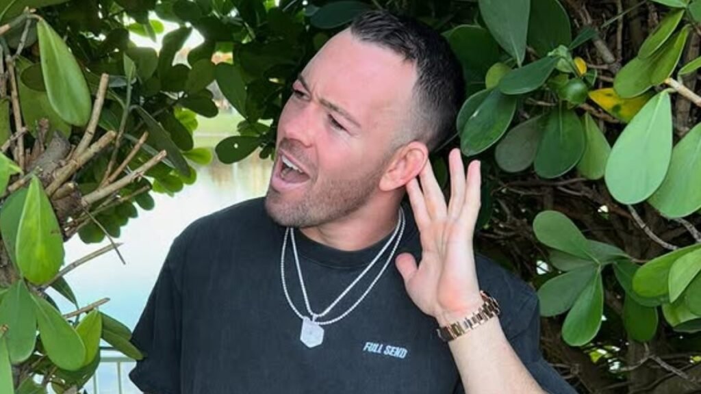 Colby Covington, luchador de MMA, con expresión dramática y mano en la oreja, "escuchando" al público entre plantas.
