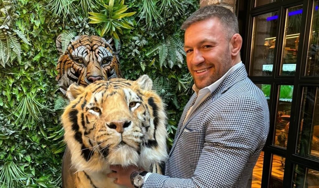 Conor McGregor sonríe con blazer de pata de gallo junto a un tigre realista y otro tallado en una pared verde.
