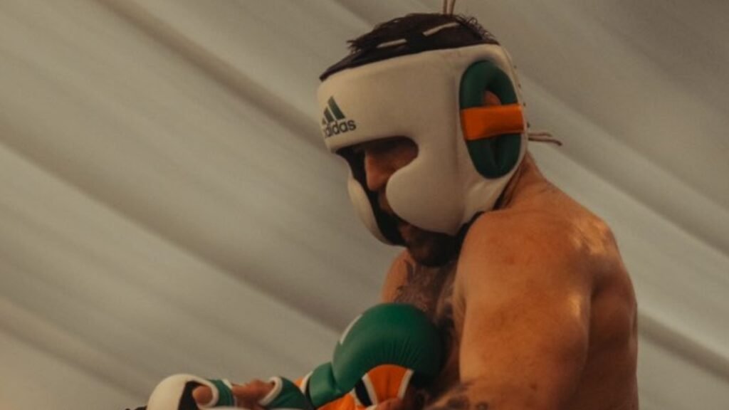 Boxeador concentrado entrenando, lleva casco protector blanco Adidas y guantes de boxeo verdes y naranjas.