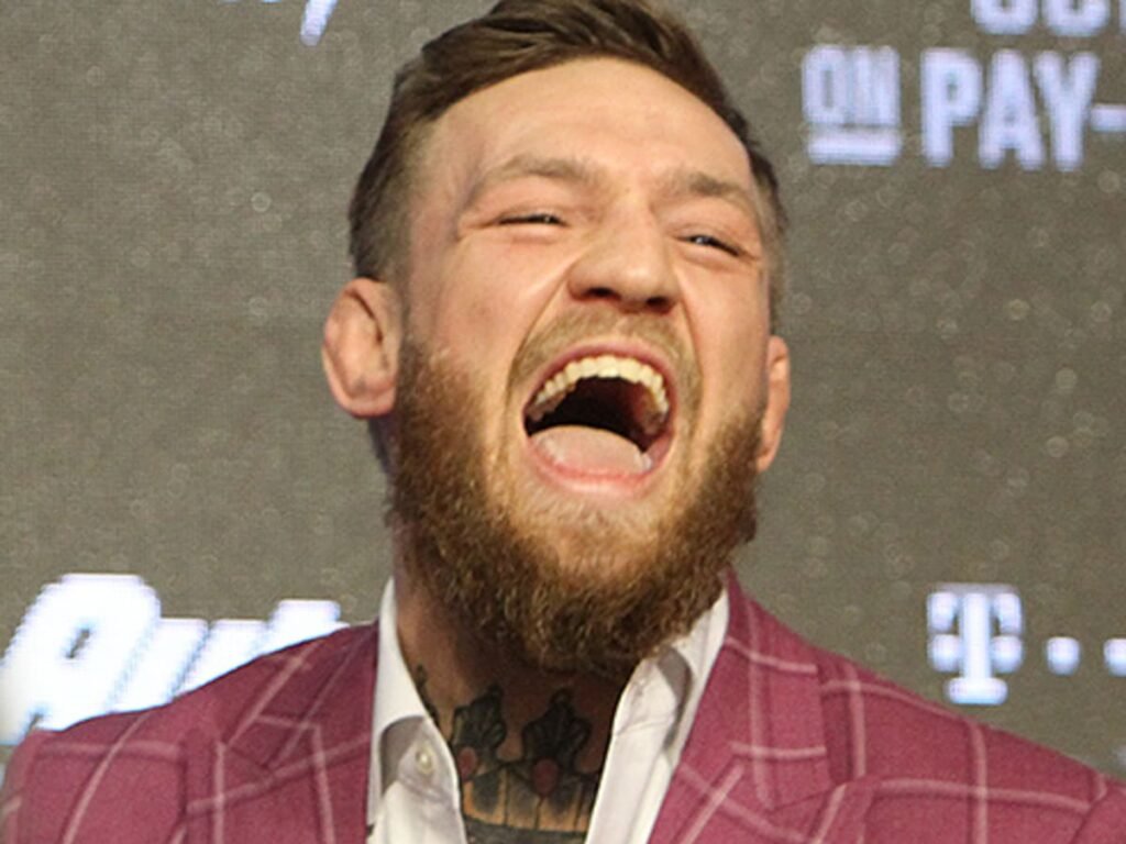 Conor McGregor, luchador barbudo, con traje granate, ríe a carcajadas o grita en una conferencia.