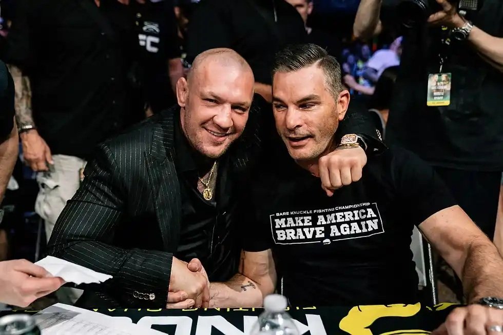 Conor McGregor y David Feldman, presidente de BKFC, posan sonrientes. Feldman le abraza y su camiseta dice "Make America Brave Again".