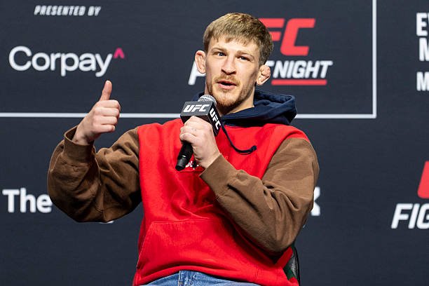 Luchador UFC con hoodie rojo/marrón, pulgar arriba. Sostiene micrófono UFC en conferencia de prensa, fondo de logos.