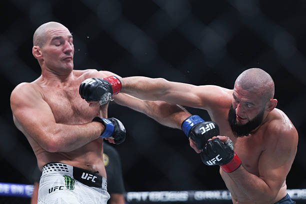 Combate UFC: Un luchador barbudo conecta un puñetazo a su rival calvo; gotas de sudor vuelan por el impacto.