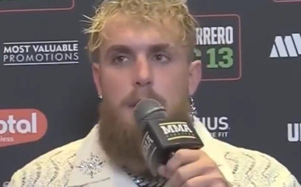 Jake Paul, boxeador e influencer, habla por micrófono MMA. Pelo rubio y barba, camisa clara. Fondo oscuro con logos de promociones.