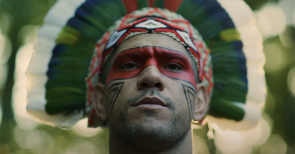 Hombre indígena con tocado de plumas (verde, blanco, azul) y pintura facial roja con diseños negros. Mirada intensa en bosque.