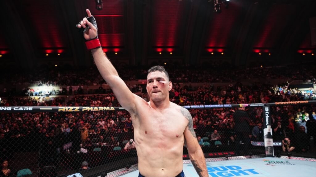 Chris Weidman, peleador de MMA, celebra su victoria en la jaula. Brazo alzado, corte en el ojo, ante una multitud.