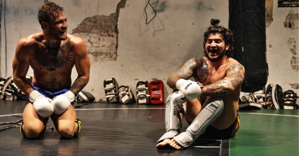 Conor McGregor y otro luchador, sonriendo y sudorosos, descansan en el tatami tras entrenar. Guantes, espinilleras y tatuajes.