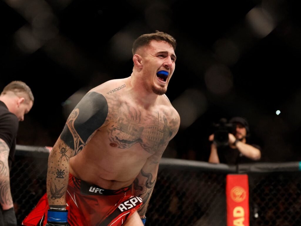 Luchador de MMA Tom Aspinall con bucal azul grita eufórico en el octágono, mostrando tatuajes y sus shorts de UFC.