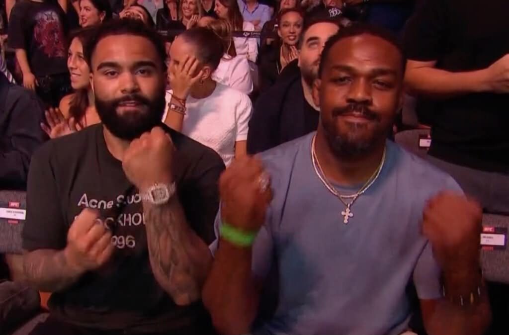 Jon Jones y Gable Steveson, puños cerrados y sonriendo, disfrutando de la acción en el UFC 327.