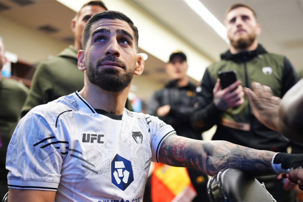 Ilia Topuria, luchador UFC, vendándose la mano en el backstage. Mirada concentrada, camiseta blanca y brazos tatuados.