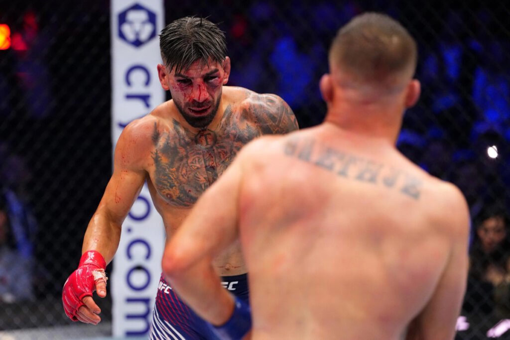 Ilia Topuria en la jaula UFC, el rostro ensangrentado y expresión tensa. De espaldas, borroso, se ve a Justin Gaethje.